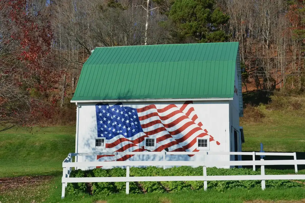Small white barn with an American flag mural behind a white fence